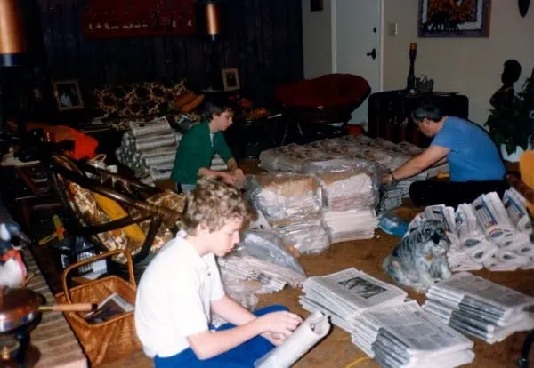 Three guys sitting on carpet putting Sunday newspapers together for paper route